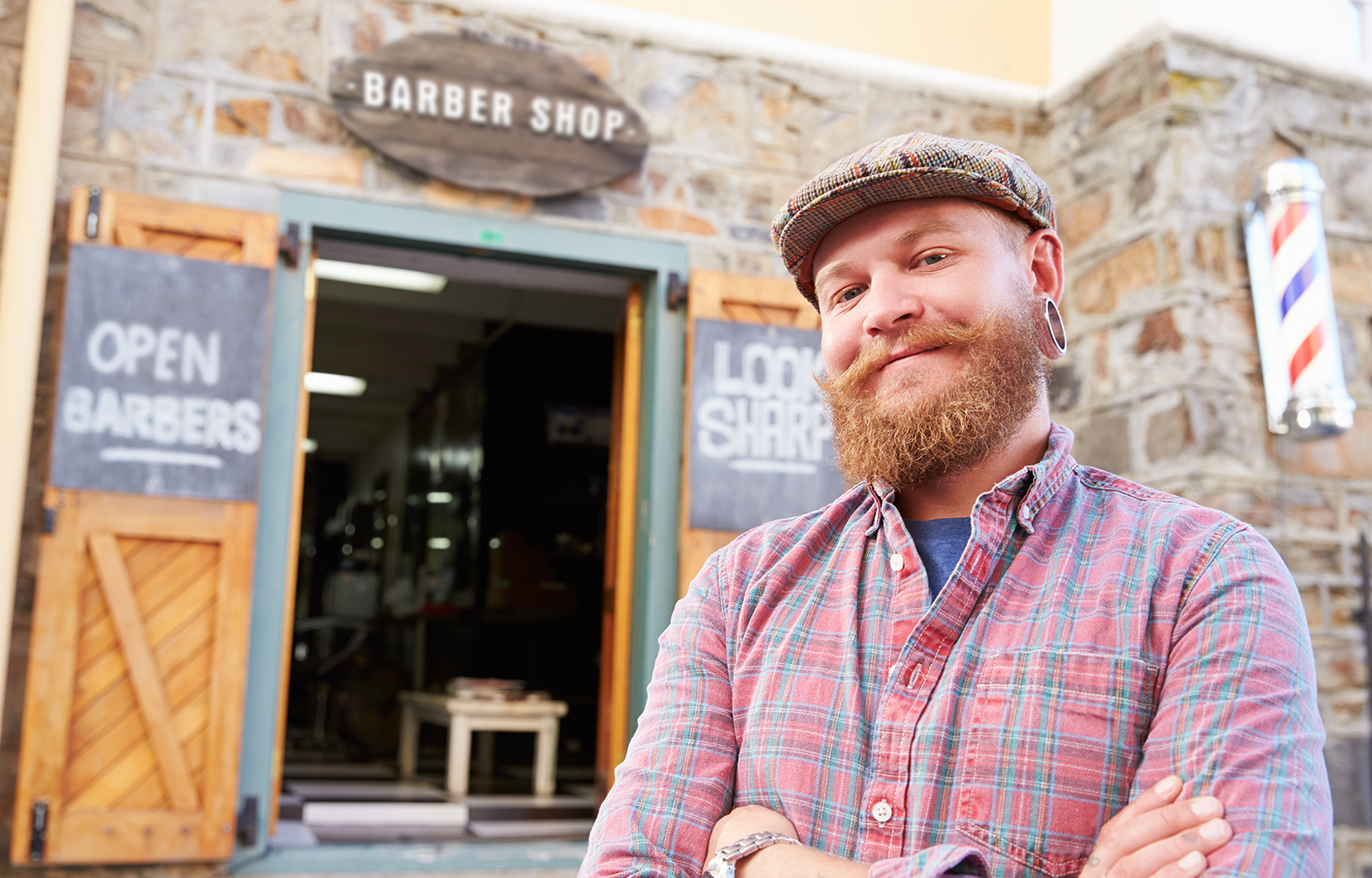 Hipster barber is smiling at camera, standing outside of his shoe doors.